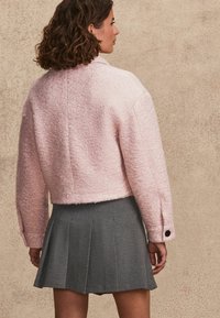 Světle růžová zkrácená bunda s texturovaným povrchem a velkým límcem, doplněná šedou plisovanou sukní s pasem a sklady.