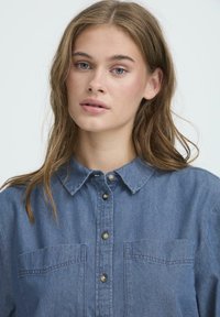 Blaues Denim-Hemd mit Knopfverschluss, Kragen, zwei Brusttaschen und einem leicht lockeren Schnitt mit sichtbaren Nähdetails.