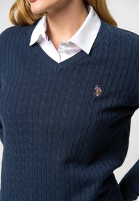 Marineblauwe trui met kabelgebreide textuur, met V-hals ontwerp, gedragen over een wit overhemd met kraag. Bevat een klein geborduurd logo op de borst.