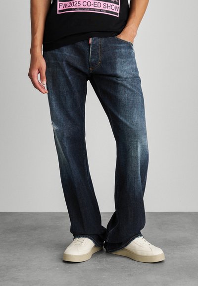 Jeans din denim de culoare albastru închis, cu o croială lejeră, prezentând ușoare decolorări și o peticire uzată. Poartă adidași albi, pe un fundal neutru.