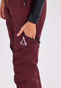 Pantaloni da esterno color bordeaux con tasca con zip, caratterizzati da un logo bianco e una texture liscia. Presentano un pannello laterale di contrasto nero.