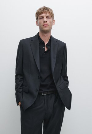 Completo scuro su misura con giacca a un petto, pantaloni abbinati e una polo nera. Tessuto liscio con hardware minimale.