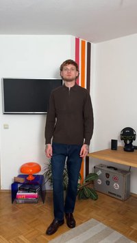 Rudas užtrauktukas megztinis su griovelių tekstūra, tamsiai mėlynos džinsai ir rudi odiniai batai. Stovi kambaryje su spalvotomis sienų juostomis ir dekoru.