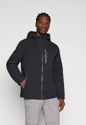 Homme portant une veste à capuche noire avec fermeture éclair à l'avant et poches zippées, associée à un pantalon cargo gris clair, posant devant un fond uni.