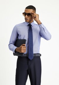 Lichtblauwe shirt met kleine patronen, donkerblauwe stropdas, gestructureerde donkere tas en donkere broek. Vergezeld van een paar bruine zonnebrillen.