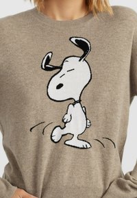 Pull en tricot beige avec un motif noir et blanc de Snoopy marchant, col rond et poignets côtelés. Texture douce, design décontracté.