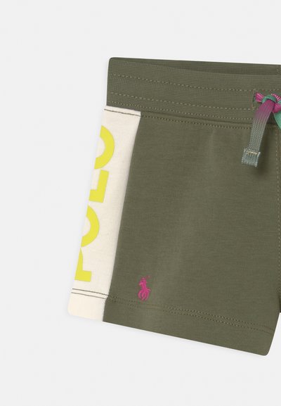 Calções verdes-azeitona com um painel bege apresentando um grande texto amarelo "POLO". Pequeno logótipo bordado rosa na bainha e cordão multicolorido.