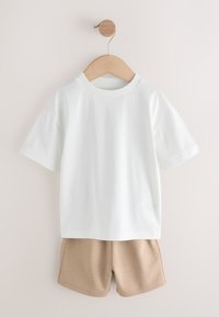 T-shirt blanc à manches courtes et shorts beige suspendus sur un cintre en bois, devant un mur clair.