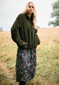 Olijfgroen pluche fleece jack met een hoge kraag, gecombineerd met een maxi-jurk met een donker bloemenpatroon in groene en roze tinten.