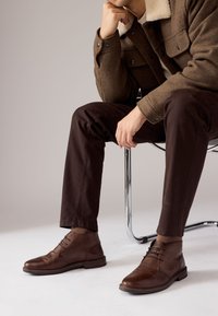 Hnedé kožené spoločenské topánky s hladkým povrchom, okrúhlou špičkou a tenkými šnúrkami, nosené s tmavohnedými nohavicami. Na neutrálom pozadí.