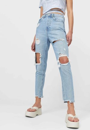 Hellblaue zerrissene Jeans mit großen Rissen an Oberschenkeln und Knien, getragen mit weißen Plateausandalen und einem kurzen Oberteil mit Wickelbändern.