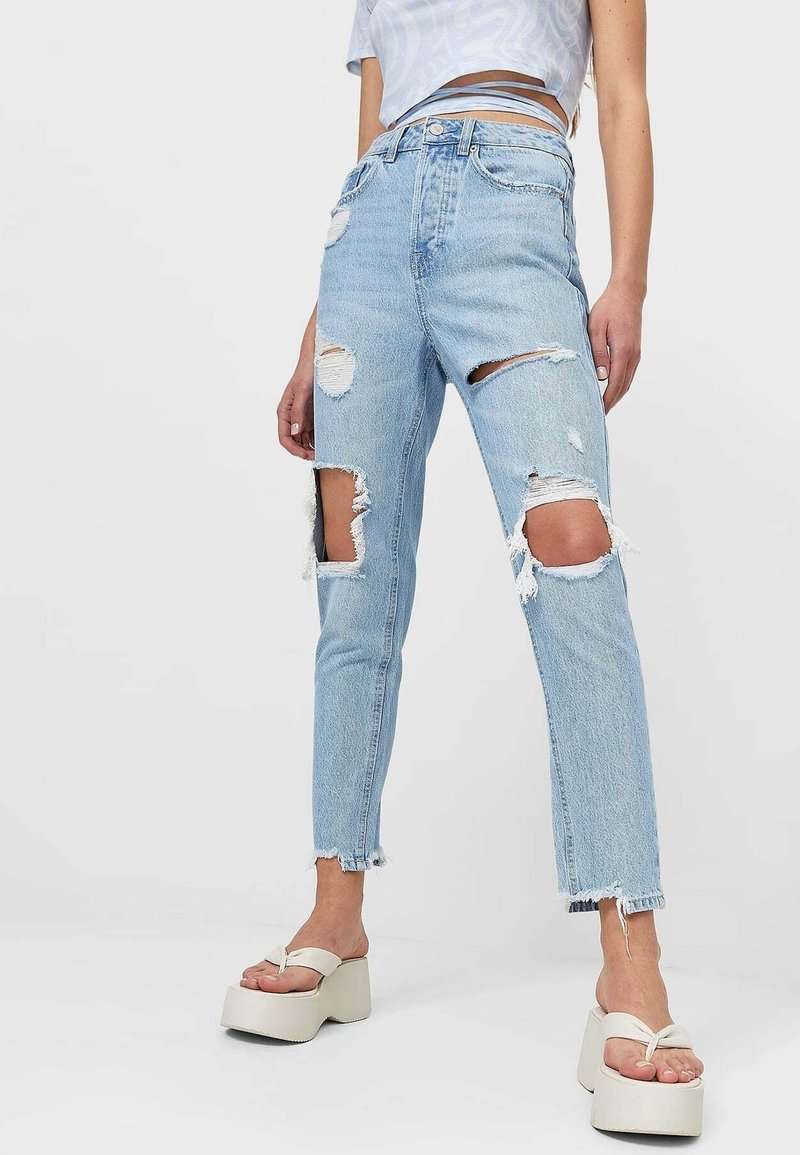 Jean déchiré bleu clair avec de grandes déchirures aux cuisses et aux genoux, porté avec des sandales à plateforme blanches et un haut court à liens enveloppants.