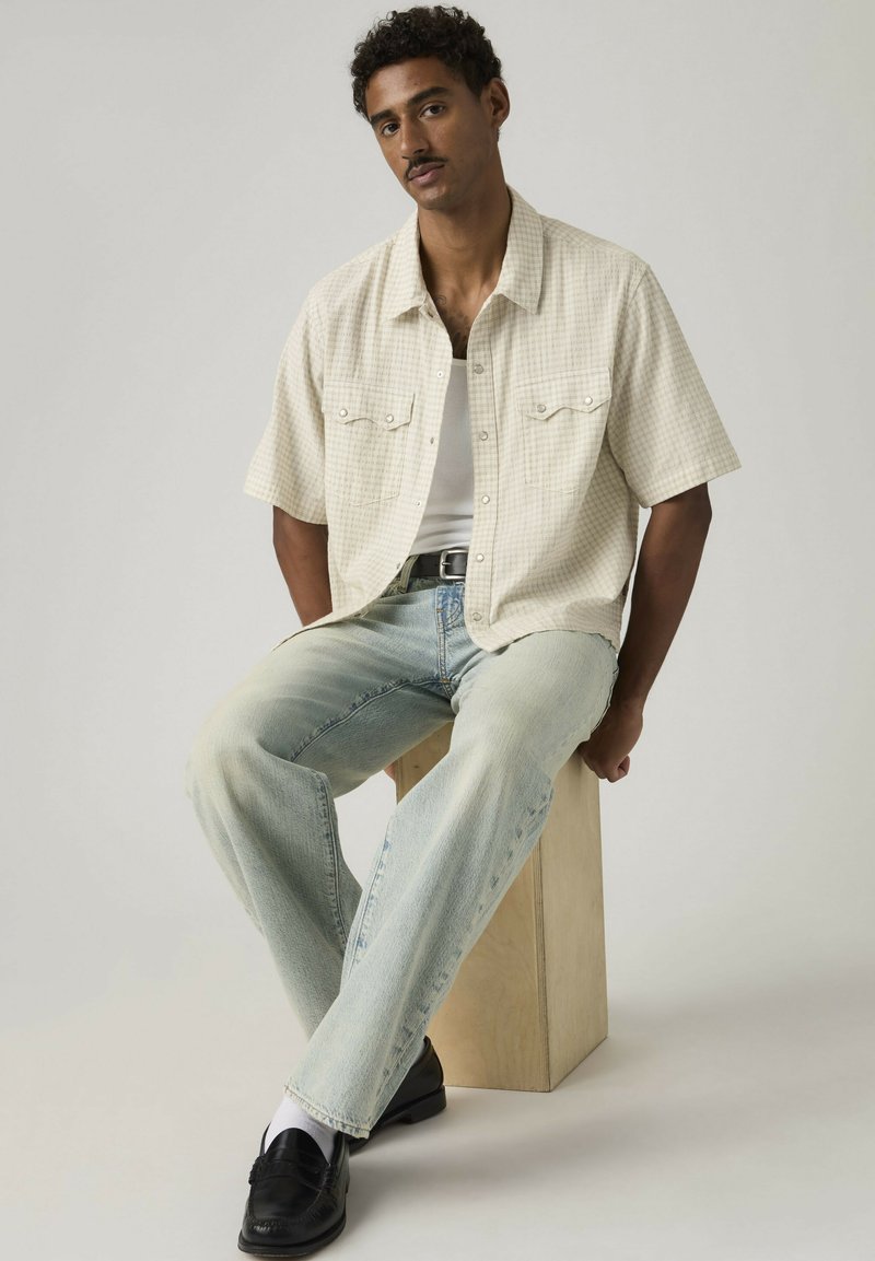 Hombre con cabello corto y rizado sentado sobre un bloque de madera, lleva una camisa de manga corta a cuadros clara, camiseta interior blanca, vaqueros desgastados y zapatos negros.