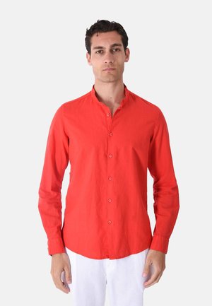 Uomo che indossa una camicia rossa a maniche lunghe con bottoni e colletto alla mandarino e pantaloni bianchi, in piedi davanti a uno sfondo chiaro e uniforme.