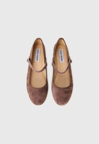 Pari avokkaista ruskeasta mokasta, joissa on nilkkahihnat ja beige pohjallinen, jota koristaa "Steve Madden" -merkintä valkoisella taustalla.