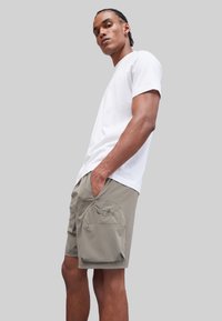 Witte t-shirt met korte mouwen, gecombineerd met kaki shorts met een grote zijzak en geborduurd logo, tegen een grijze achtergrond.