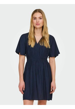 Kvinde med langt blondt hår iført en navyblå kortærmet kjole med V-hals og indsnøret talje, stående mod en ensfarvet lys baggrund.