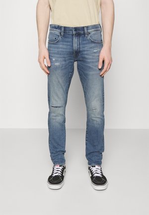 Mężczyzna noszący jasnoniebieskie, postrzępione, obcisłe jeansy i czarne trampki z białymi sznurówkami, stojący na jednolitym białym tle.