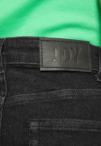 Black denim jeans featuring a textured leather label embossed with "JDY" on the back waistband. The fabric has a subtle sheen.