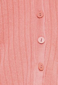 Růžový žebrovaný svetr s pěti kulatými knoflíky vpředu, který vystavuje strukturovaný pletený vzor a měkkou, lehkou tkaninu.