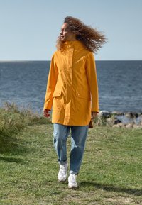 Gul vattentät jacka med hög krage, två stora framfickor, kombinerad med ljusblå jeans och vita sneakers, på gräsbevuxen mark vid vatten.