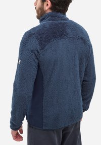Mężczyzna w ciemnoniebieskiej polarowej kurtce z wysokim kołnierzem, stojący tyłem do kamery na białym tle.