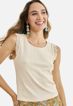 Mujer sonriente con cabello largo, vestida con un top sin mangas crema con ribete de encaje y pantalones con estampado floral, complementada con pendientes colgantes coloridos.
