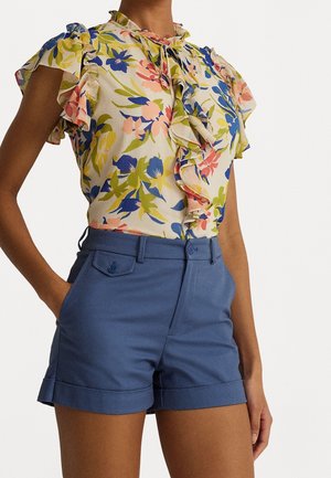 Floral blouse with ruffled sleeves and collar paired with high-waist blue cuffed shorts, hand in pocket, against a white background.