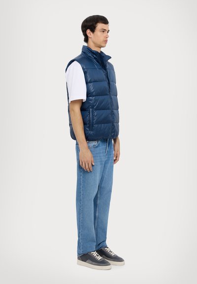 Navy blue puffer vest with high collar and front zipper, worn over a white t-shirt and paired with light blue jeans and dark sneakers.