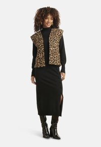 Gilet con stampa leopardata sopra un maglione nero a maniche lunghe e un vestito midi con spacchi laterali e bottoni. Stivali neri alti fino al ginocchio completano il look.