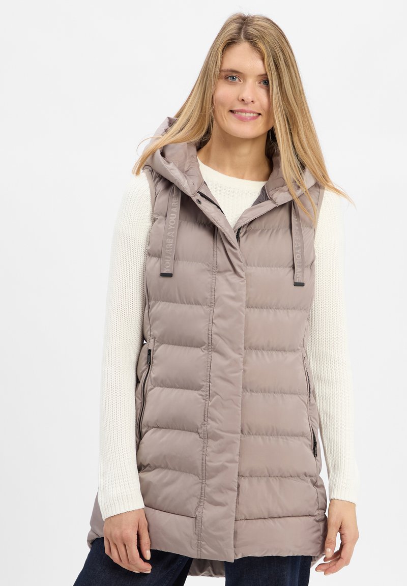 Gepolsterte Weste in hellem Taupe mit Kapuze, seitlichen Reißverschlusstaschen und Frontreißverschluss, über einem weißen Strickpullover getragen.