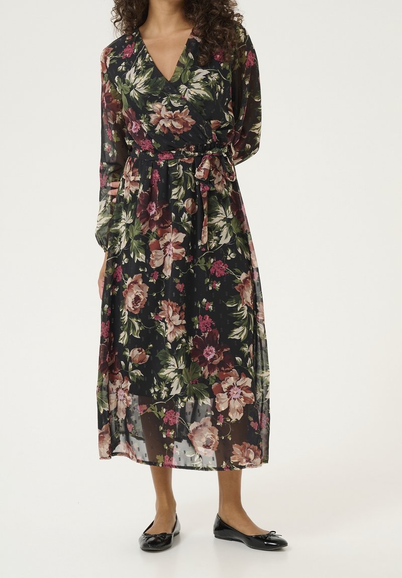 Femme portant une robe midi noire à manches longues à motif floral avec une ceinture nouée à la taille et des ballerines noires, posant devant un fond uni.
