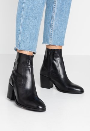 Paar schwarze Leder-Stiefeletten mit Blockabsatz, getragen zu hellblauen Jeans mit ausgefranstem Saum auf weißem Boden.