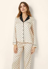 Polka dot pajama set in beige satin with black accents. Features long sleeves, button-up front, and a pocket on the top.