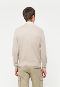 Ung kvinne med ryggen til, iført en beige strikkegenser over en hvit skjorte og lyse khaki cargo-bukser, mot en enkel hvit bakgrunn.