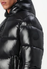 Black puffer jacket with a shiny finish, featuring a hood, zippered pockets, and a round logo patch on the sleeve.