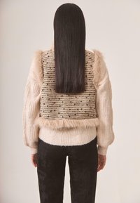 Jersey de punto con una textura de trenzas beige, que presenta un chaleco texturizado con acentos negros y un borde de piel sintética en la parte inferior y los hombros.
