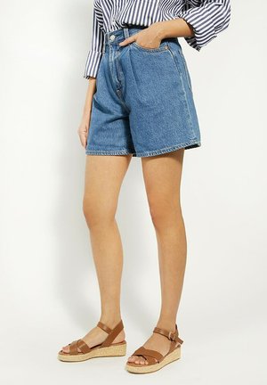 Person wearing high-waisted blue denim shorts, striped long-sleeve shirt, and brown wedge sandals with ankle straps, standing against a white background.