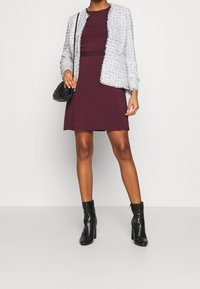 Veste en tweed grise aux bords effilochés portée sur une robe bordeaux. Bottines noires à talons. Petit sac à main noir tenu dans la main gauche.