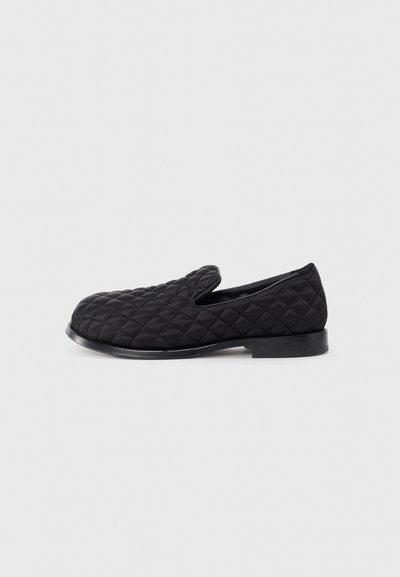 Sort quiltet slip-on sko med et struktureret stofoverdel, afrundet tå og en flad sort sål, der viser en diamantmønster syning.