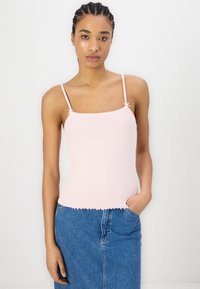 Lys rosa camiskjorte med spagettistropper, ribbet tekstur og bølget kant. Kombinert med et denimskjørt, poserer modellen mot en nøytral bakgrunn.