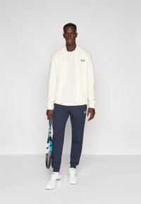 Ανδρική αθλητική εμφάνιση με μπεζ ζακέτα με φερμουάρ, λευκό μπλουζάκι, μπλε σκούρο φόρμα με λογότυπο και λευκά αθλητικά παπούτσια, κρατώντας ρακέτα τένις.