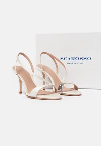 Witte sandalen met hoge hak en dunne bandjes, waarvan één met een zilverkleurig accent, geplaatst voor een witte doos met het opschrift "SCAROSSO Made in Italy."