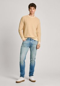 Suéter de punto beige claro con cuello redondo, combinado con jeans azules deslavados y zapatillas blancas con suelas marrones. Diseño minimalista.