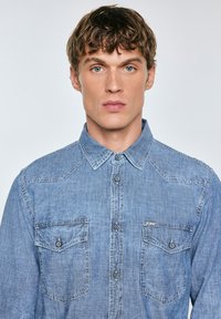 Camisa de denim en un tono azul claro, con cierre de botones, cuello clásico y dos bolsillos en el pecho con solapas abotonadas. Tejido texturizado.