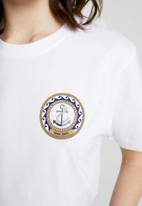 Hvid bomulds T-shirt med et cirkulært grafik af en anker, reb og blå bølger, ledsaget af teksten "MOIN, MOIN" forneden.