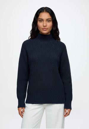 Femme aux longs cheveux foncés portant un pull en maille côtelée bleu marine et un pantalon blanc, debout devant un fond clair uni.