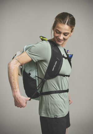 Woman adjusting straps on Ortovox hiking backpack, wearing light green shirt and black shorts, smiling and looking down.