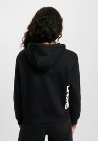 Juodas trumpas hoodie su minkštu paviršiumi, su reguliuojama kapuce ir balta logotipo spauda kairėje pusėje. Paprastas dizainas.