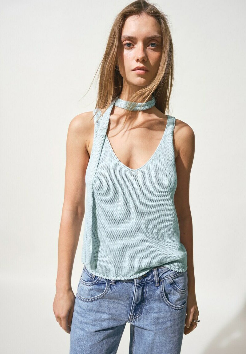 Jeune femme portant un débardeur en maille bleu clair avec un foulard assorti autour du cou et un jean en denim bleu, debout devant un fond uni.